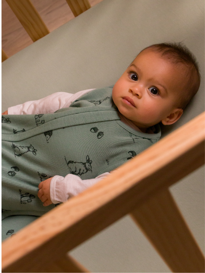 Baby lying in cot with green woodland print merino sleeping bag
