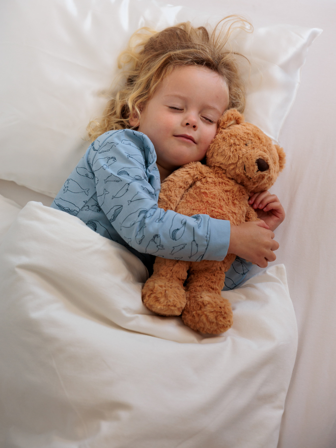 Toddler in white bdding and blue pyjamas cuddling a brown teddy bear