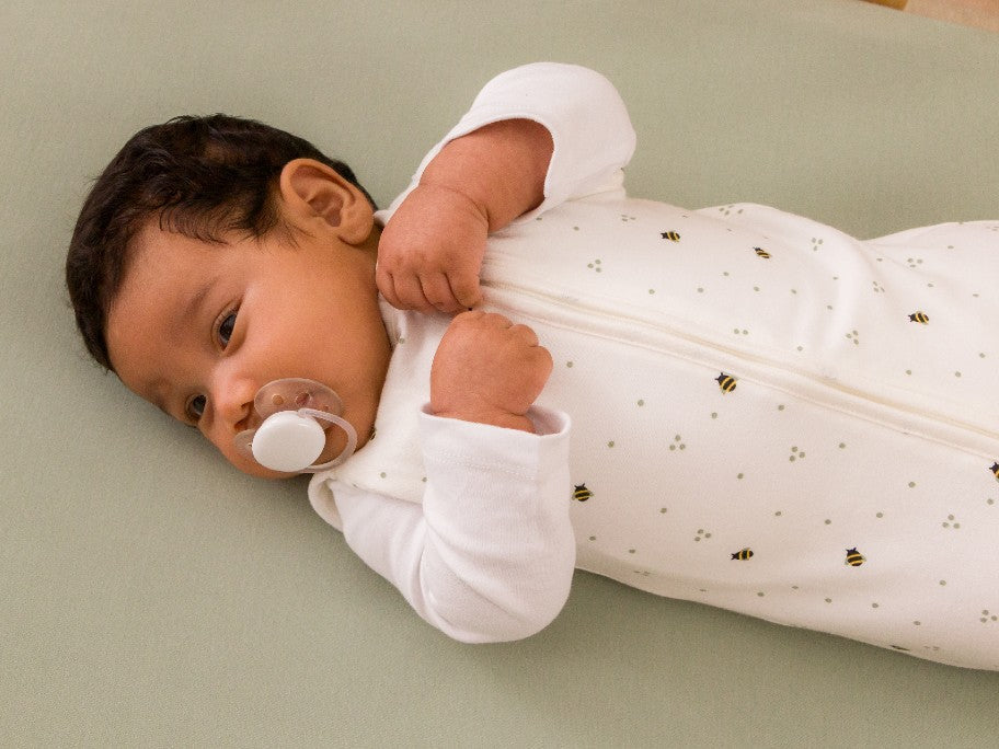 Baby in cot with dummy and wearing a cotton swaddle bag with small bee pattern