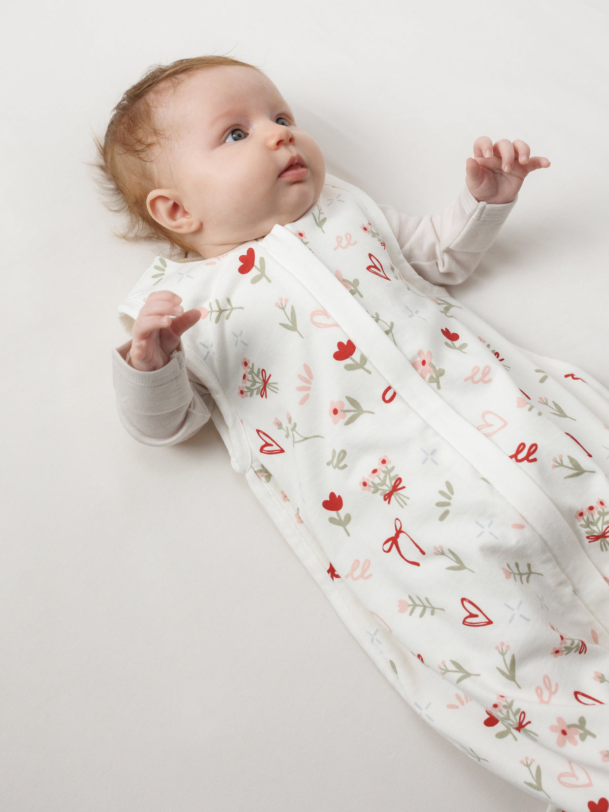 Baby llying on white sheet wearing the sleep store all seasons sleeping bag with heart, bows and flowers print