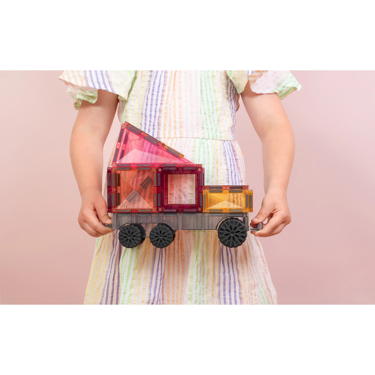 A child wearing a pastel striped dress holds a toy cargo truck made of translucent magnetic building blocks with black wheels against a soft pink background.