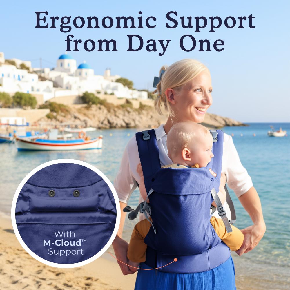 Woman carrying a baby in a blue ergonomic carrier with a scenic beach background