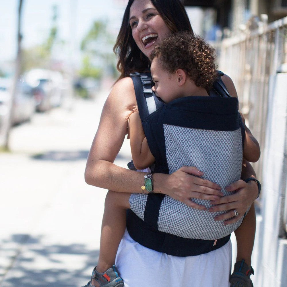 Woman carrying a 3 year old child in a mesh Beco Toddler Cool carrier on a sunny street.