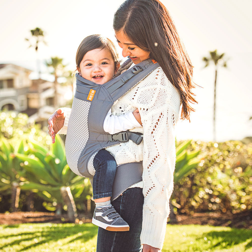 Woman carrying a two year old child in a toddler carrier outdoors with greenery in the background