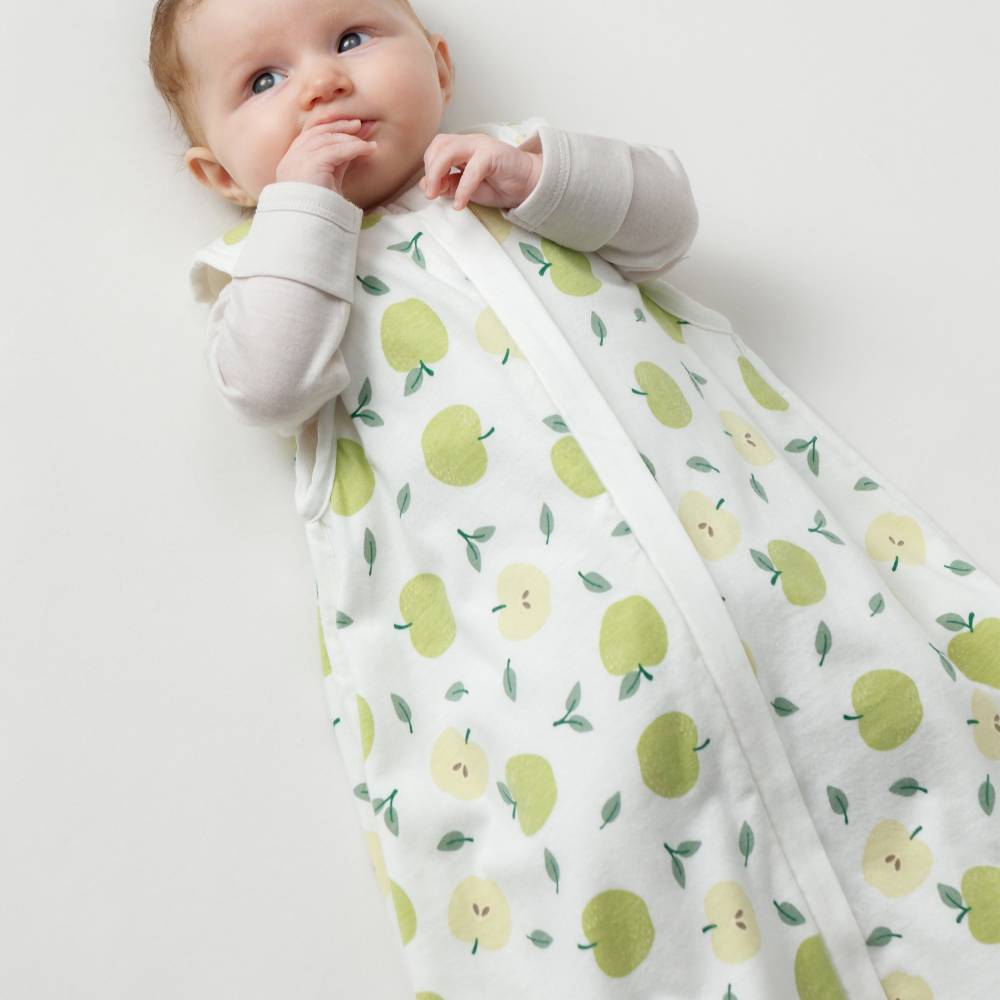 A baby wearing a white sleep sack with a green and yellow apple print, lying on a light off-white background.
