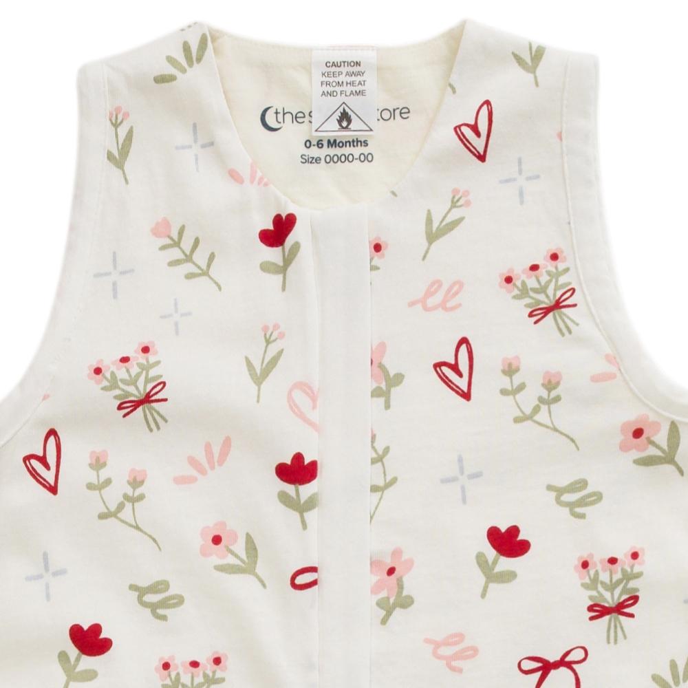 A sleeveless baby vest in off-white, decorated with a pattern of red hearts, pink flowers, green stems, and small red bows. The inner neck label shows the brand 'thesleepstore'