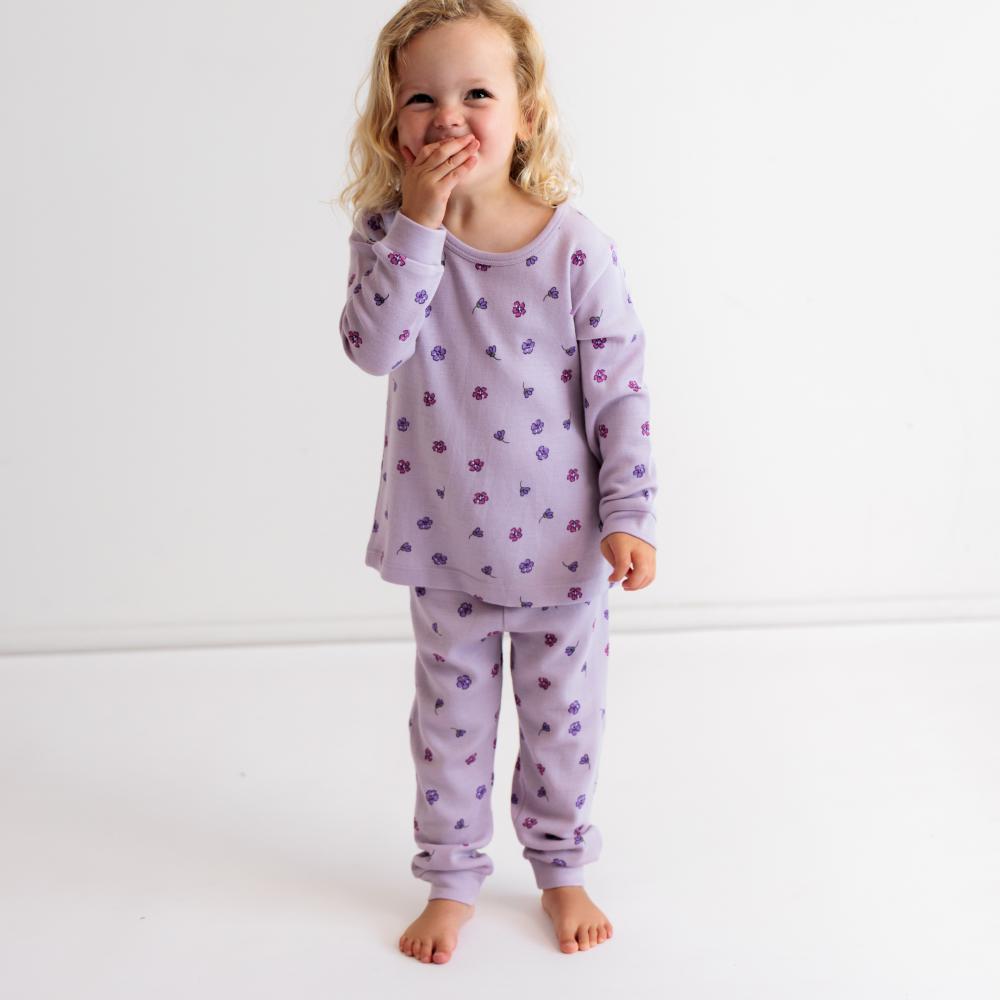 A young child wearing a light purple long-sleeve pajama set with a small scattered floral pattern, standing barefoot against a clean white studio background.