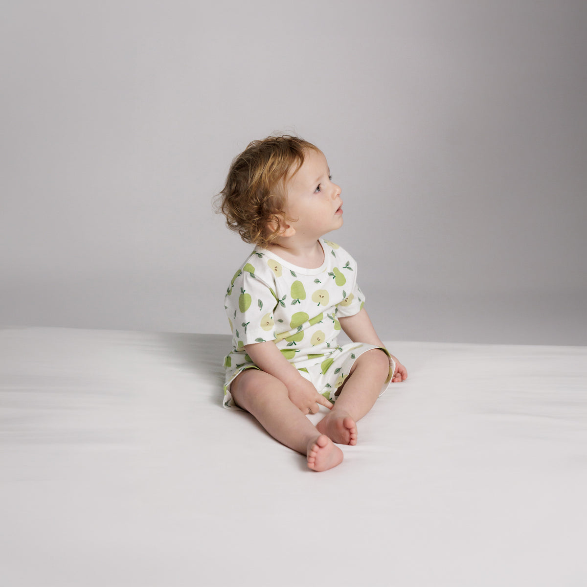 A baby sitting down wearing short sleeve cotton pyjamas with an all over apple print. 