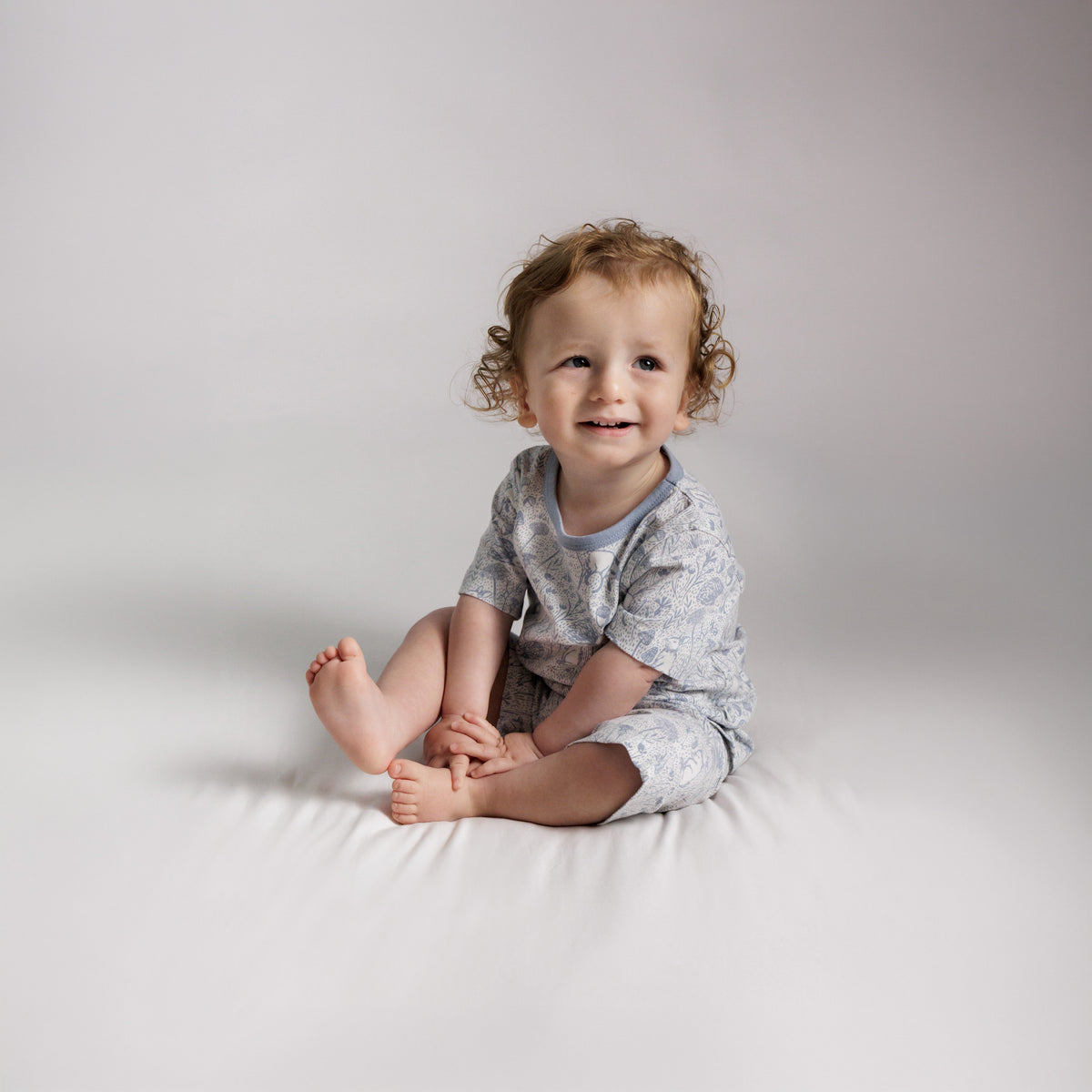 A child sitting down wearing short sleeve pjs with a blue woodlands print. 