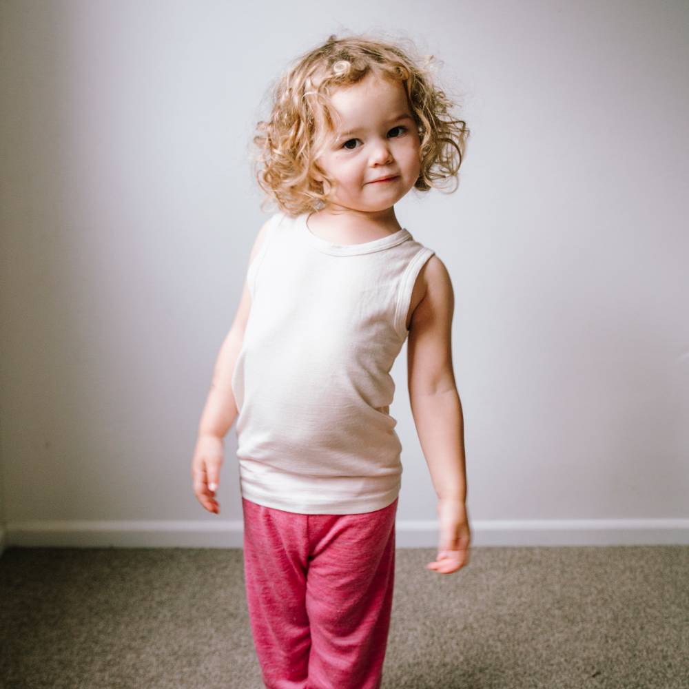 A toddler with curly blonde hair stands indoors wearing a sleeveless cream top and pink pants against a plain light gray wall and carpeted floor.
