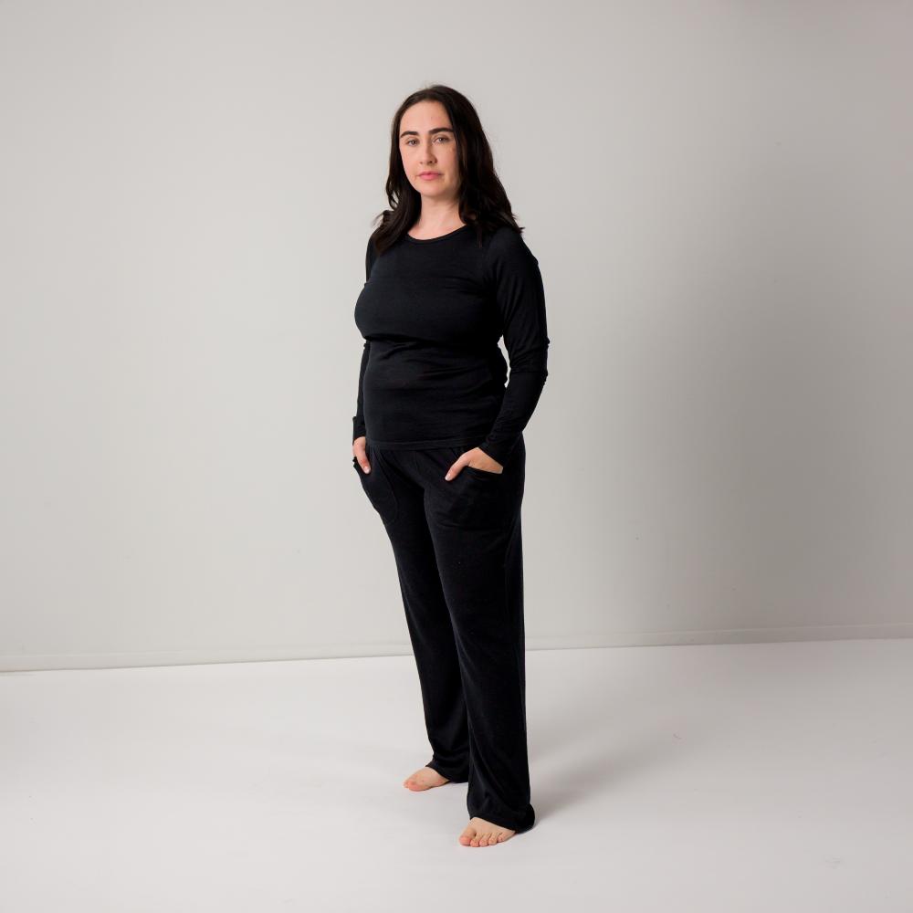 A woman modeling a black long-sleeve top and matching black pants with side pockets, standing barefoot against a plain light gray studio background.