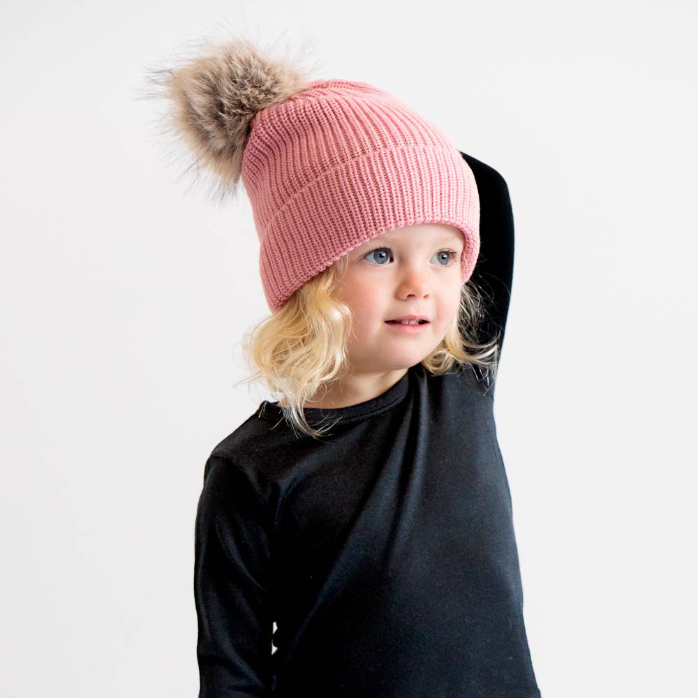 Child with blonde hair wearing a pink merino wool knit beanie with a pom-pom on a white background