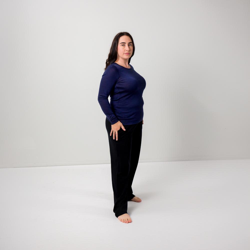 A woman stands barefoot in a studio wearing a navy blue long-sleeve top and black pants against a plain light gray background.