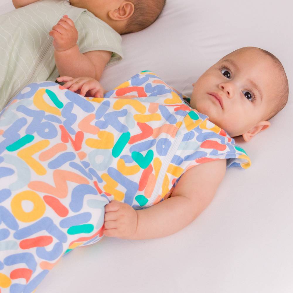 A baby lying on a white bed, wrapped in a colorful sleep sack with a playful multicolor abstract pattern. Another baby is partially visible in the background wearing a light green outfit.