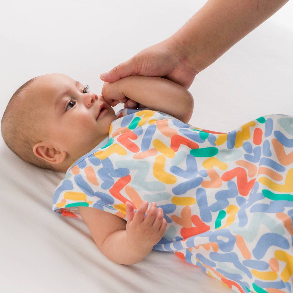 A baby lying on a white blanket while an adult hand holds the baby's hand. The baby is wearing a sleeveless sleeping bag with a colorful abstract pattern in blue, orange, yellow, and green on a white background.