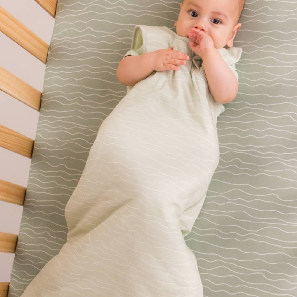 A baby lying on their back in a crib, in a pale sage green sleep sack with short sleeves. The crib mattress has a matching sage green and white wavy pattern, and the wooden slats of the crib are visible on the left.