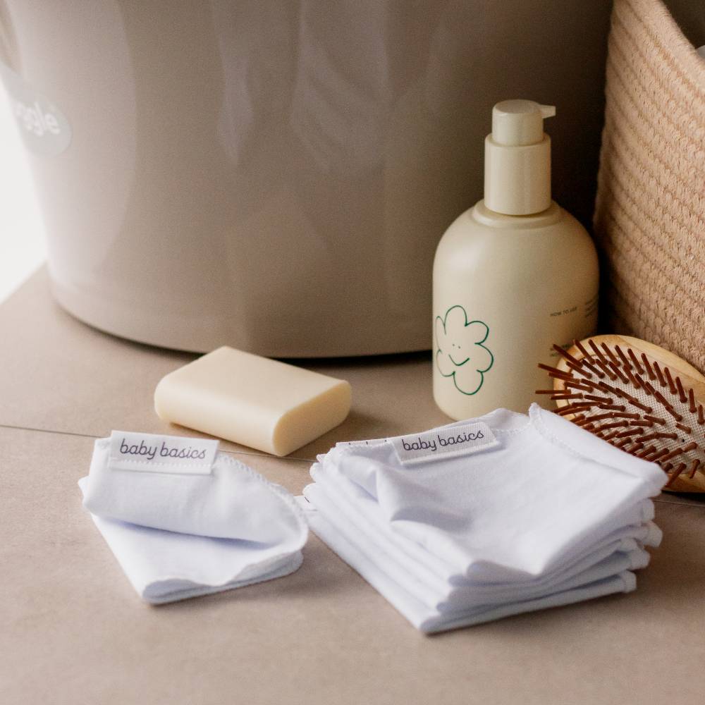 Baby care products including a bottle, soap, and wash cloths on a neutral background