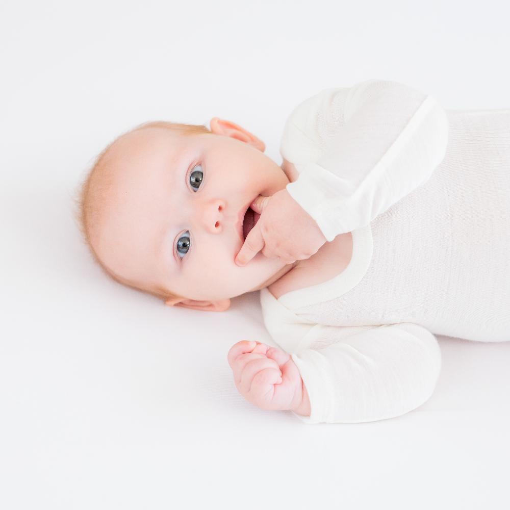 A baby lying on their side wearing a cream long-sleeve bodysuit, looking up with a hand near their mouth against a clean white background.