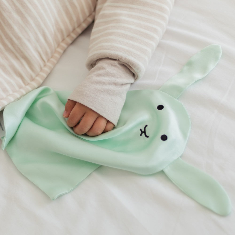 Child's hand holding a green bunny silk comforter on a white surface