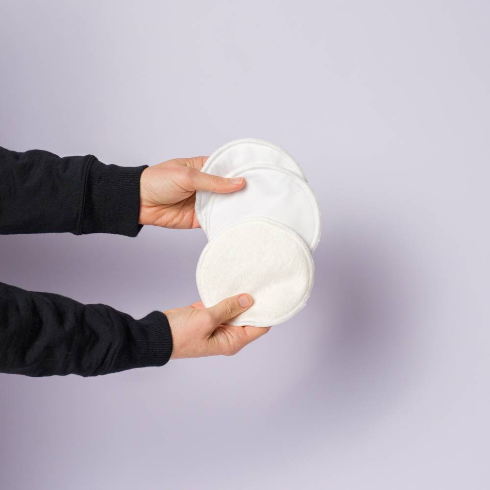 A person wearing a black long-sleeve top holds two round white fabric nursing or pumping pads against a plain light lavender background.