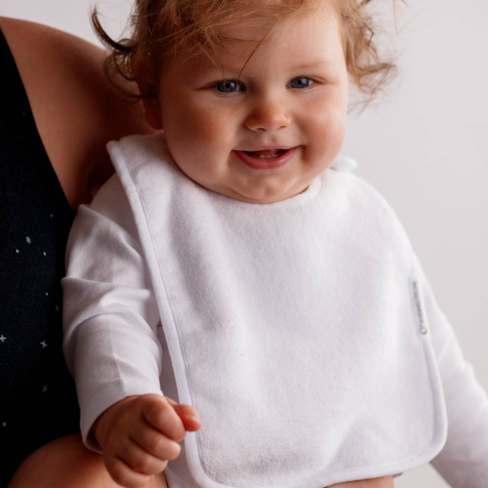 Smiling baby wearing a white cotton terry cloth classic bib with a blurred background