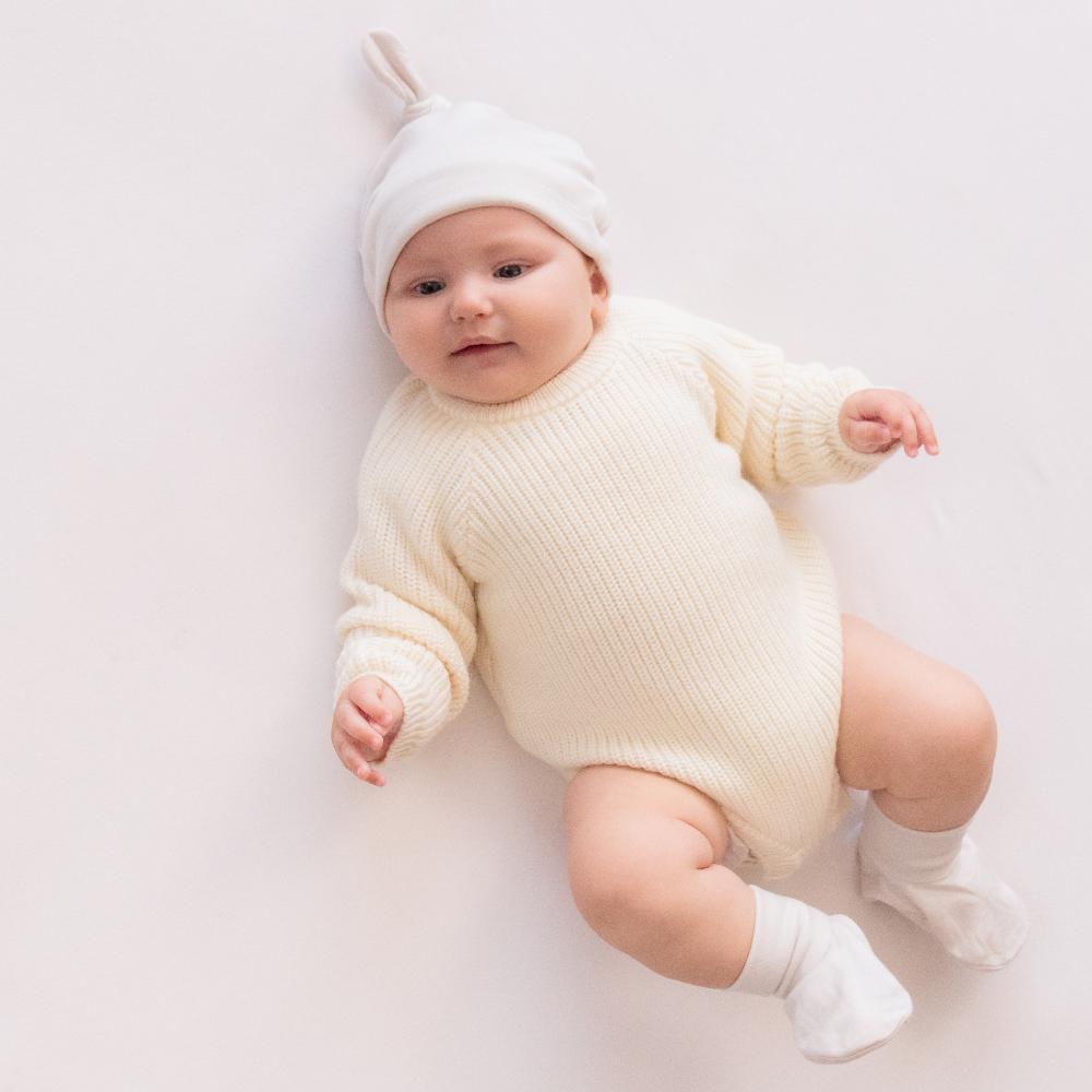 Baby wearing a white knitted merino wool bubblesuit outfit and hat on a light background