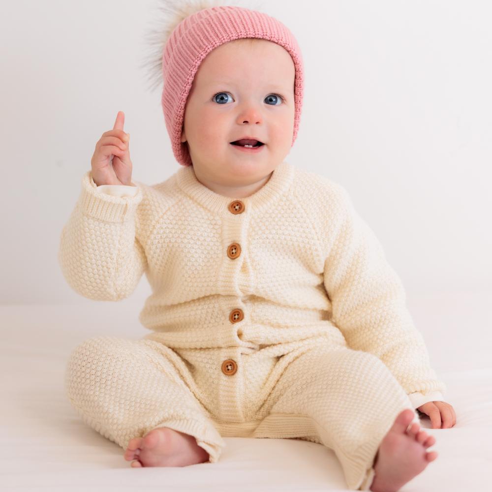 Baby wearing a cream knitted outfit with brown buttons and a pink knit hat on a white background