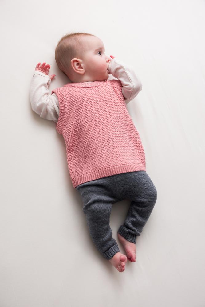 Baby wearing a pink woolen knitted vest and gray pants lying on a white surface