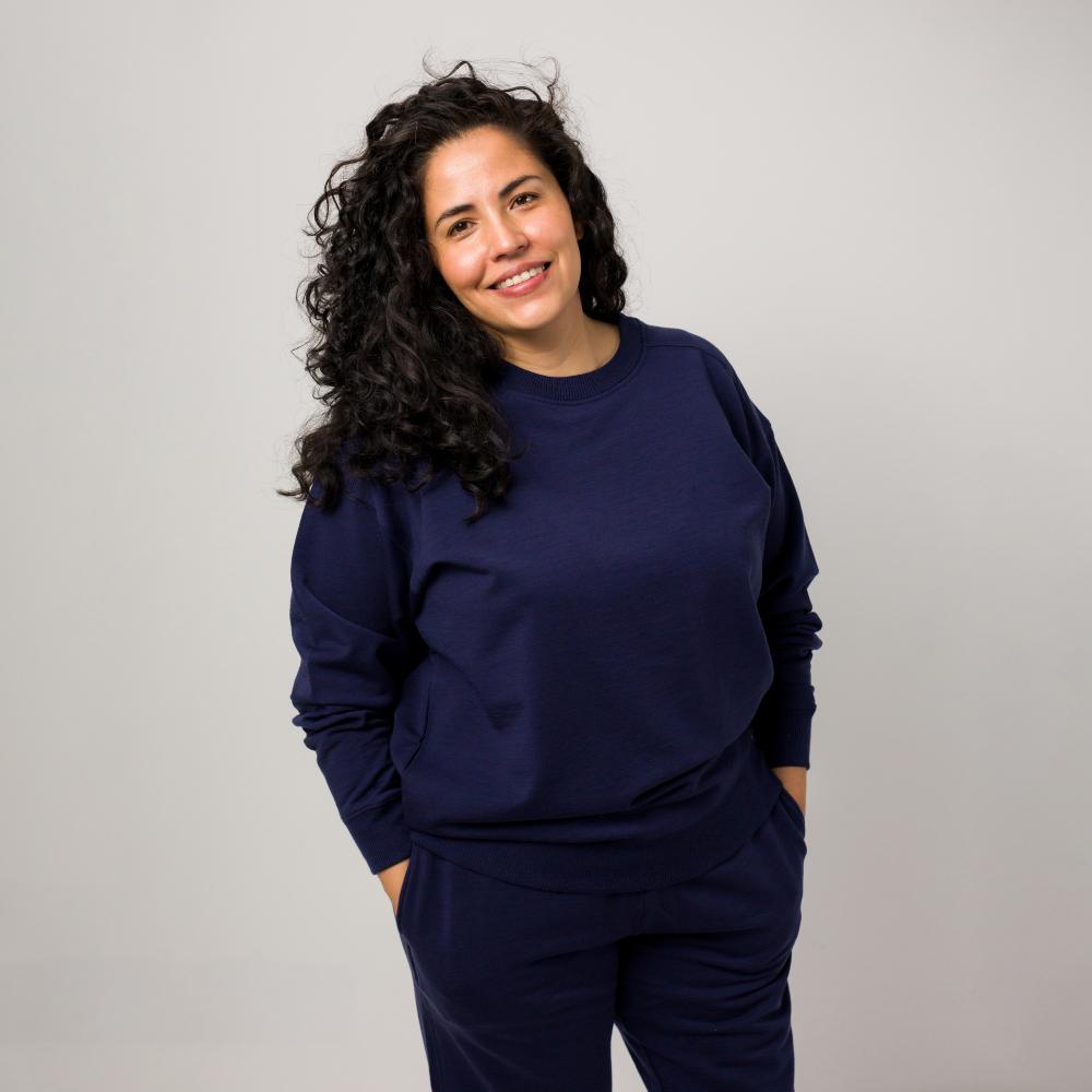 A smiling woman with curly dark hair wearing a matching navy blue merino fleece sweatshirt and sweatpants set, standing against a plain light gray studio background.