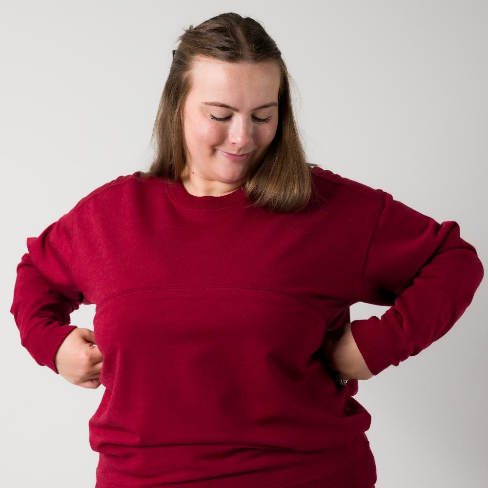 A woman wearing a dark red merino fleece crewneck nursing sweatshirt, posing with her hands on her waist against a plain light gray background.
