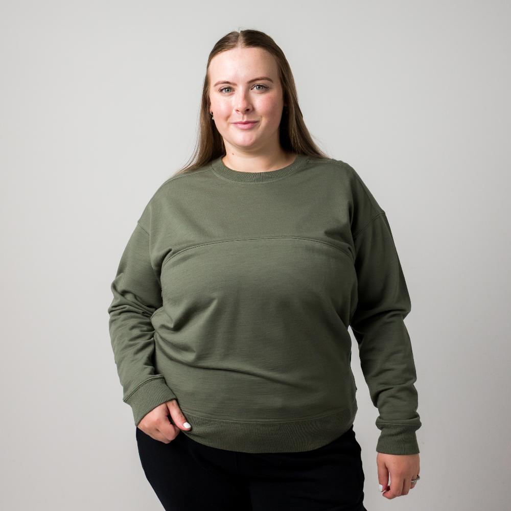 A woman modeling an olive green crewneck nursing sweatshirt with a horizontal seam detail across the chest, paired with black pants, against a plain light gray background.
