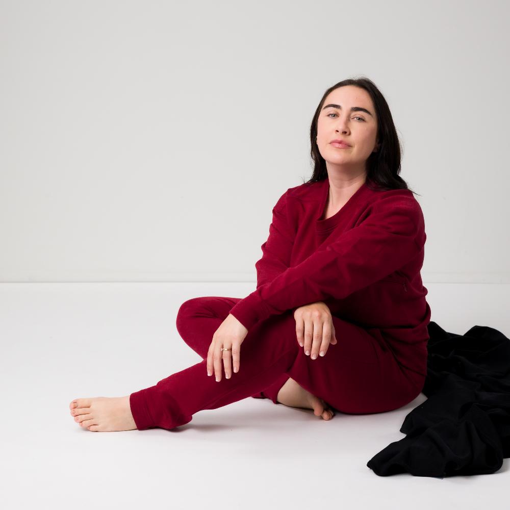 A woman sits cross-legged on a white studio floor wearing a matching deep burgundy merino fleece lounge set, with a black garment draped beside her.