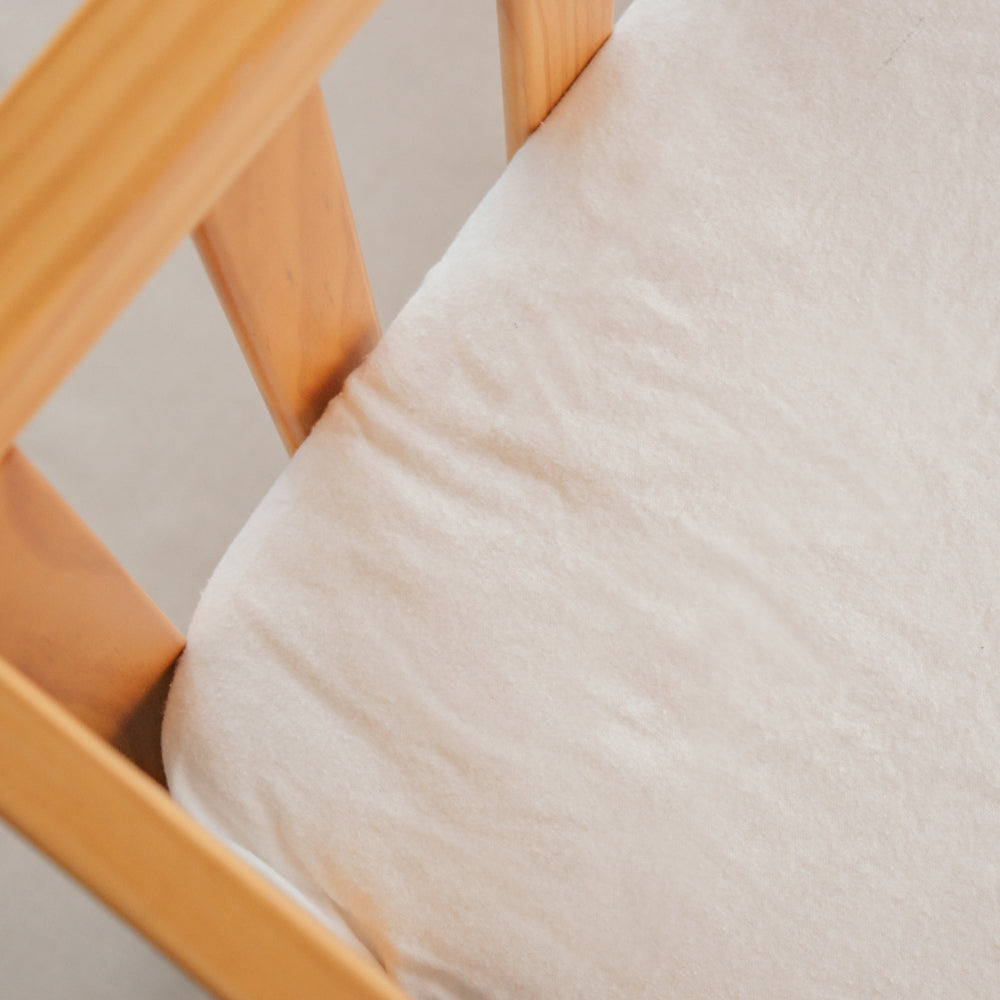 A close-up of a soft off-white or cream fitted sheet draped over the corner of a light natural wood crib or bassinet frame.
