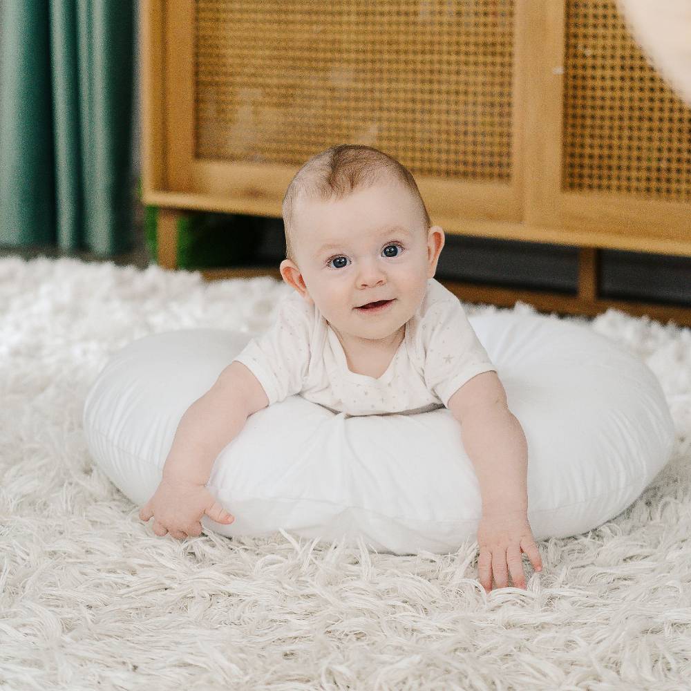 Baby lying on a white cushion on a textured floor with a wooden cabinet in the background