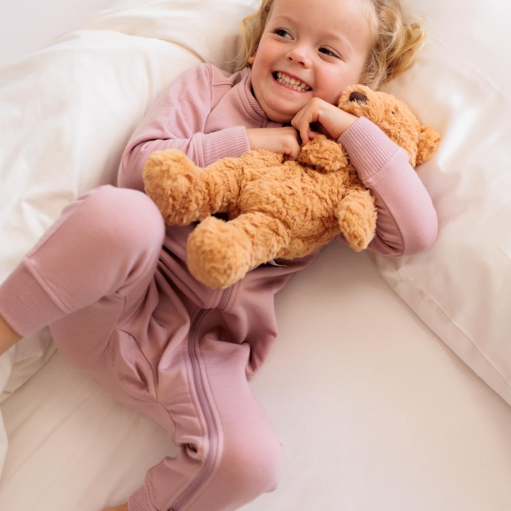 A young child wearing a dusty pink ribbed long-sleeve pajama onesie lies on a white bed, smiling while holding a brown teddy bear nightlgiht and sound machine