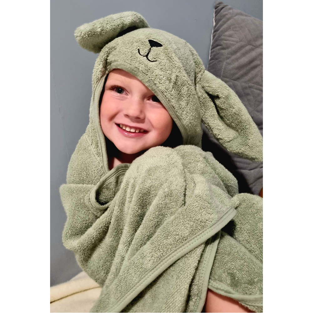 A smiling child wrapped in a plush sage green hooded towel with bunny ears and a small embroidered face, posed against a gray background.