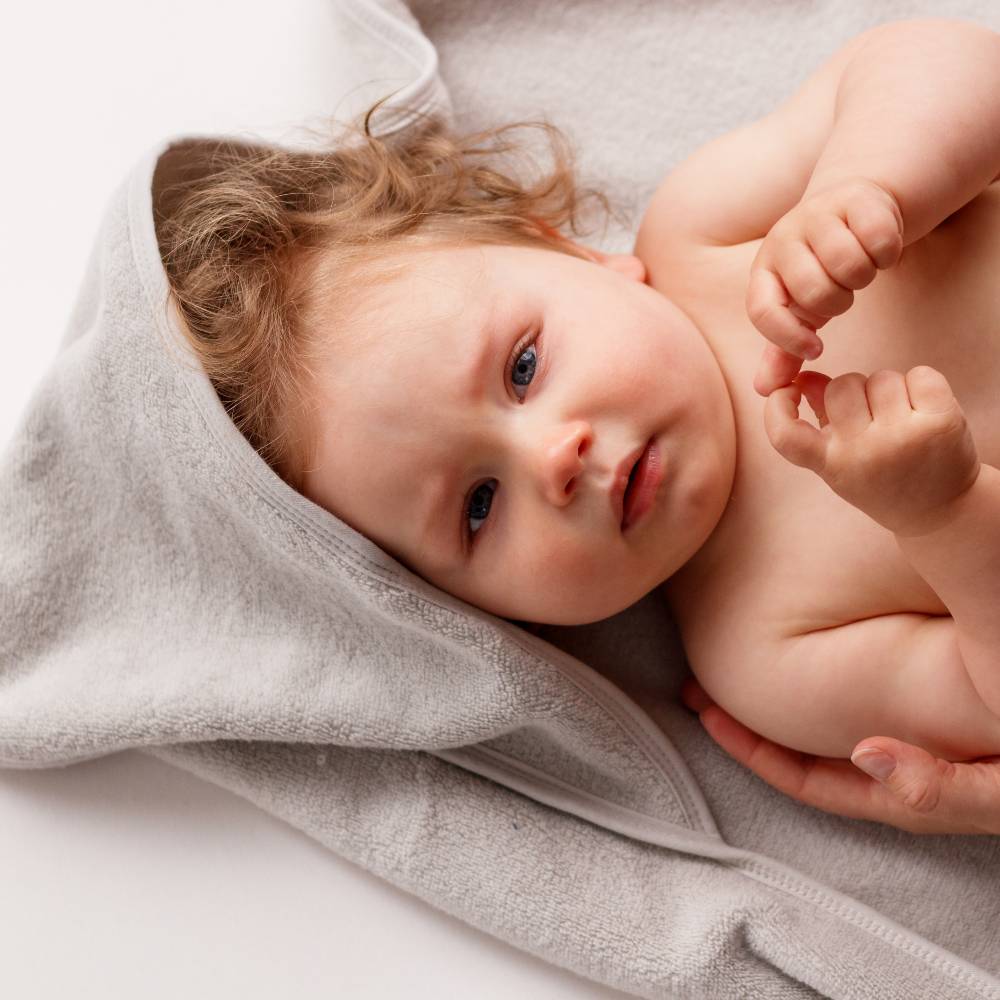 A close-up of a baby lying on their side, wrapped in a soft light gray towel. The baby has light brown hair and blue eyes, and their hands are visible near their chest.