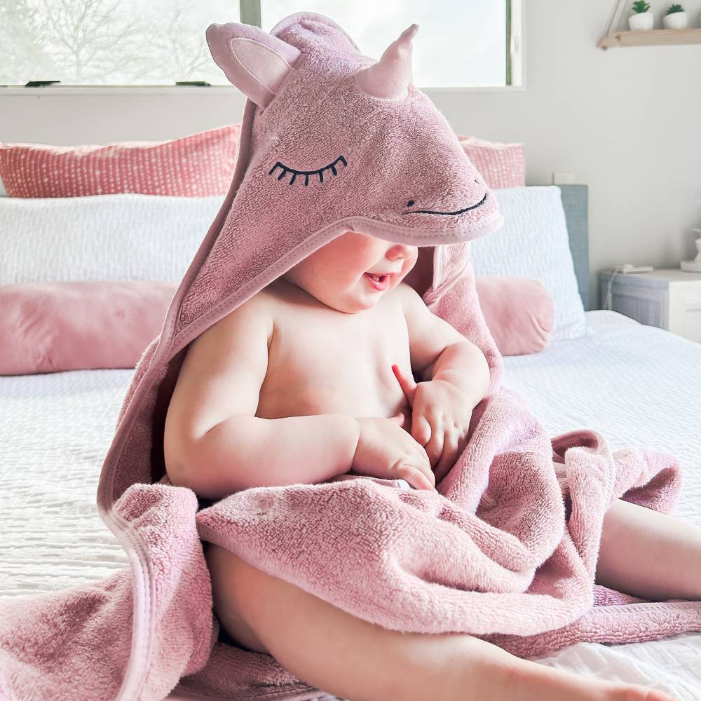 A baby sitting on a bed wrapped in a pink plush unicorn cotton terry hooded towel, with the towel's horned hood covering the child's head.