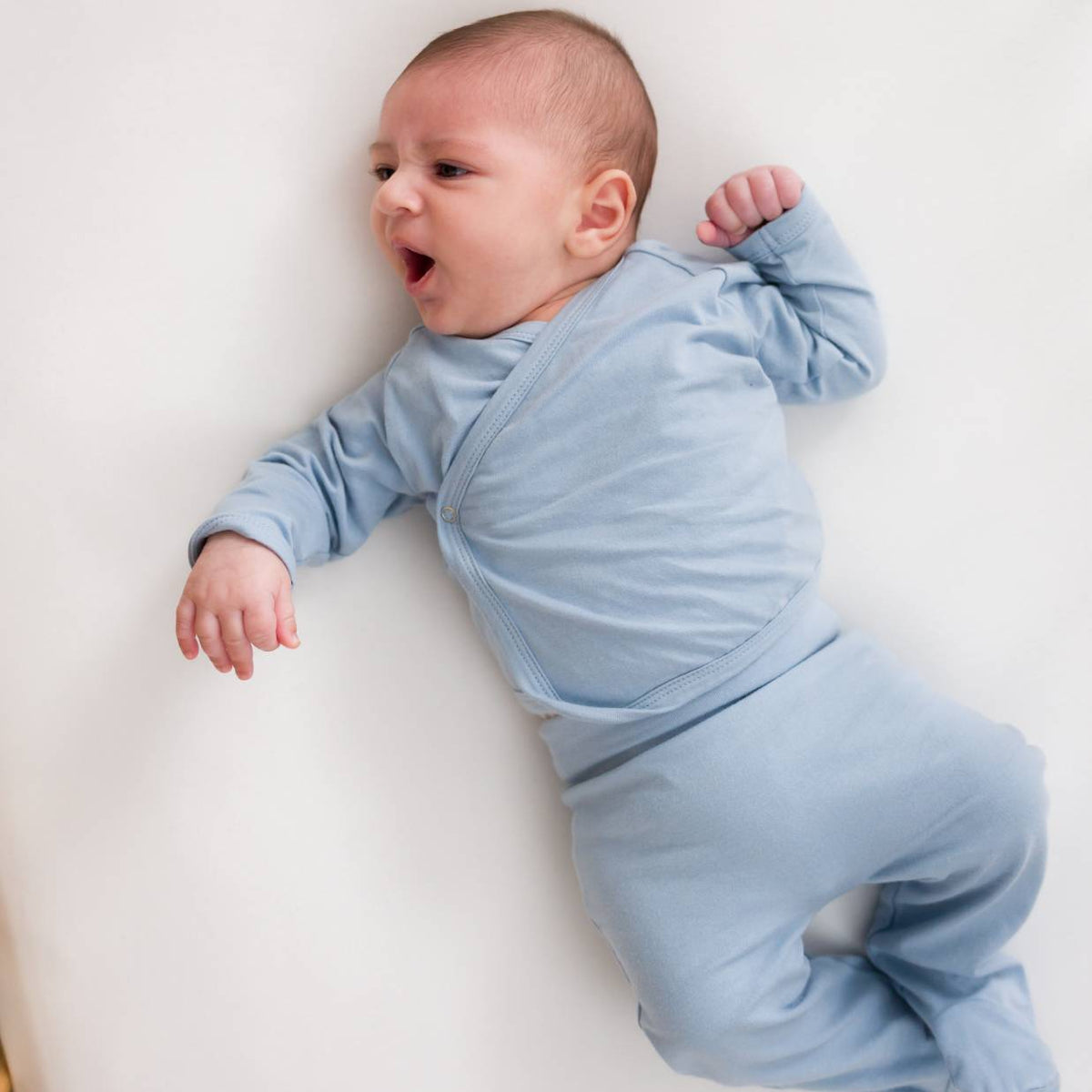 A baby lying down wearing a blue set that includes a organic cotton kimono style cardigan and pants. 