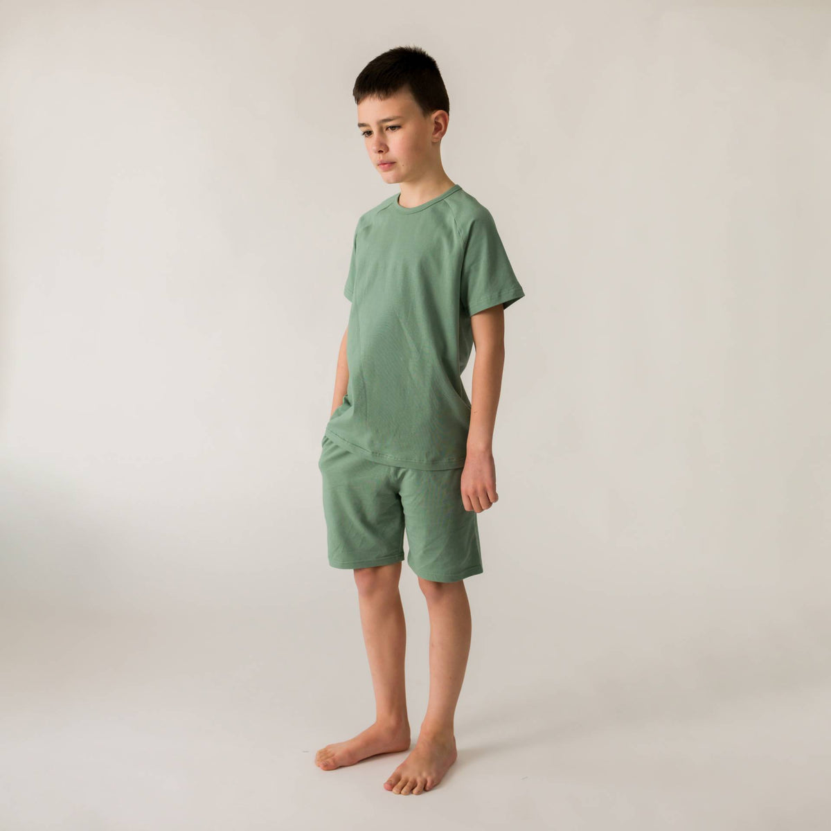 A young boy wearing fern green short sleeve pyjamas on a light grey background. 