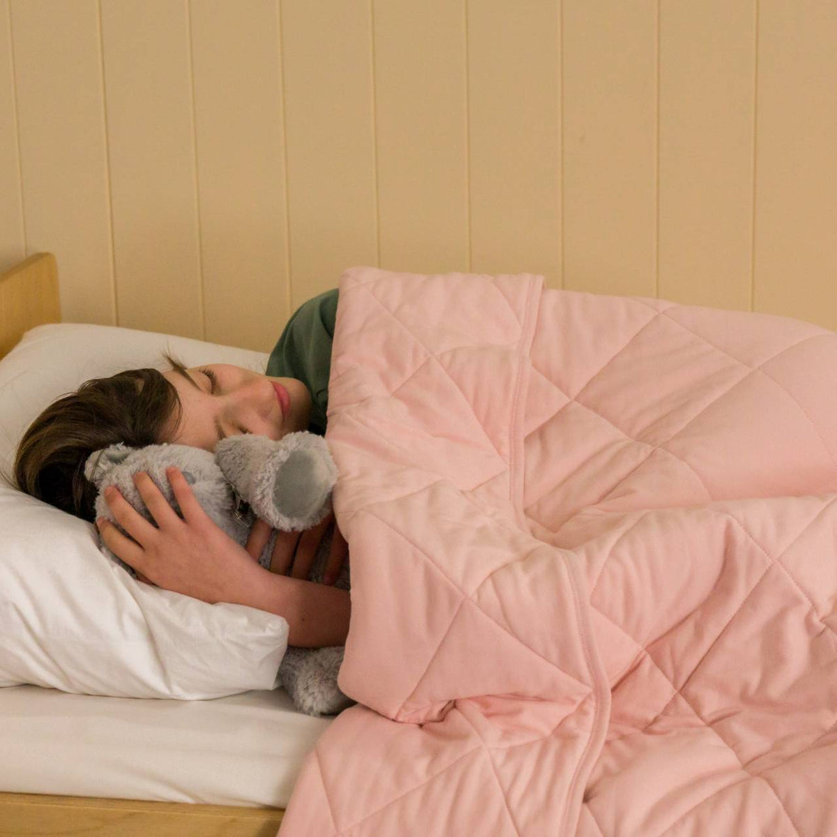 A young child cuddling a soft toy in bed under a blush pink quilted blanket. 