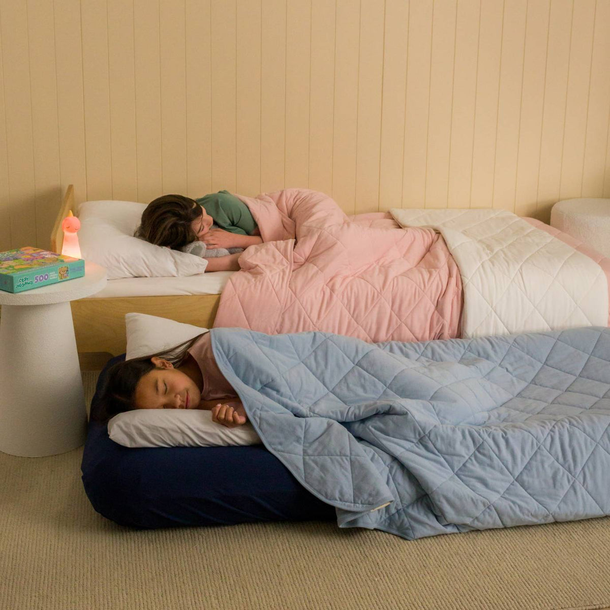 Two young children lying in bed. One with a blush pink quilted blanket and the other with a light blue quilted blanket. 