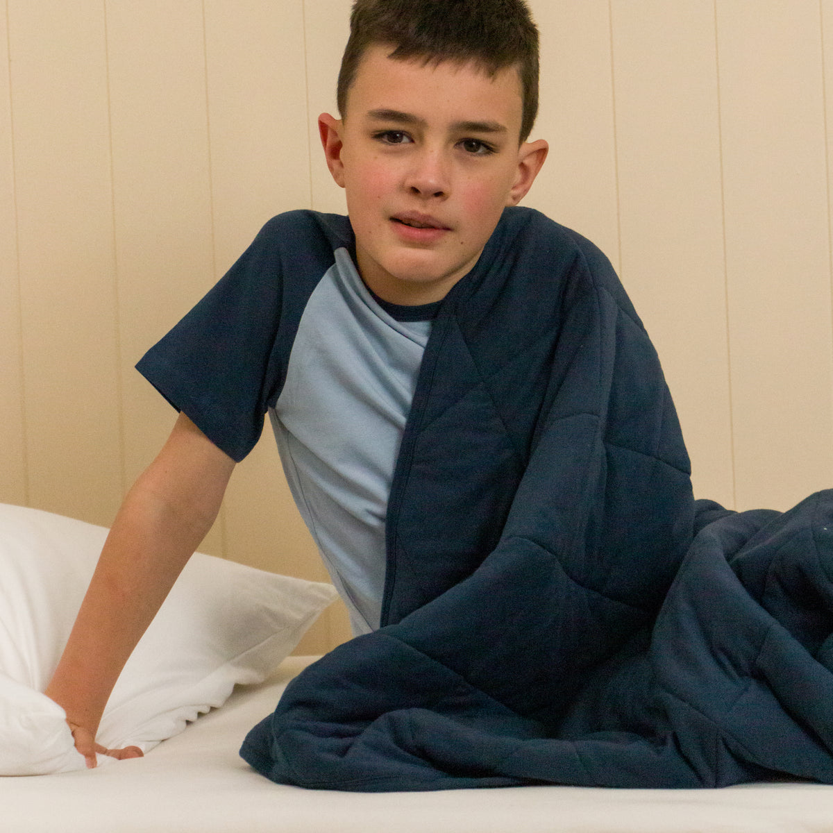 A young boy sitting up in a bed wrapped in a navy quilted blanket. 