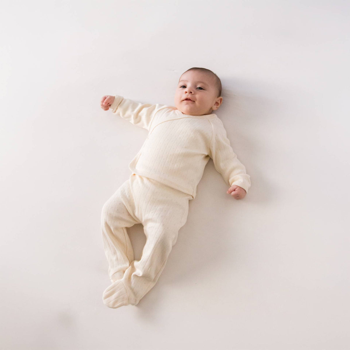 A baby lying down wearing a beige babyccino cotton pointelle kimono cardigan with matching footed pants. 