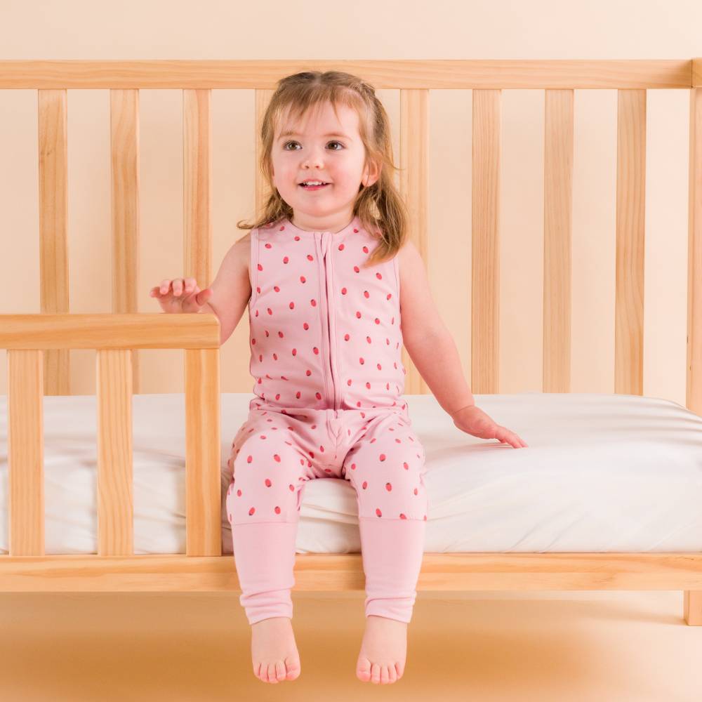 Child in pink sleepsuit sitting on a wooden crib with a plain background