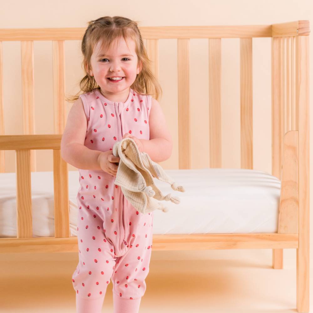 Child in pink strawberry outfit standing next to a wooden crib