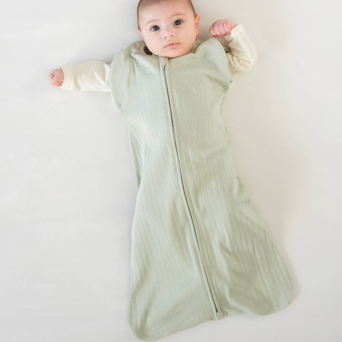 A baby lying down wearing a sage swaddle bag with arms out. 