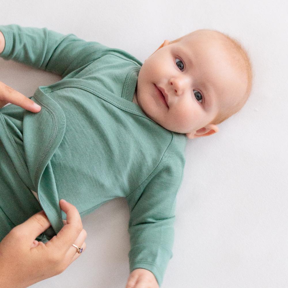 Baby lying on a white sheet wearing green jersey merino kimono style wrap cardigan and an adult hand adjusting the bottom