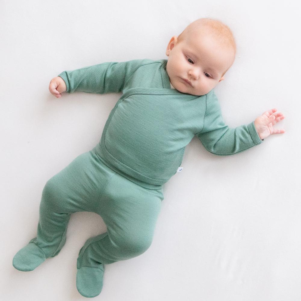 A baby lying on a white surface wearing a sage green long-sleeve kimono style bodysuit and footed pants in a jersey merino