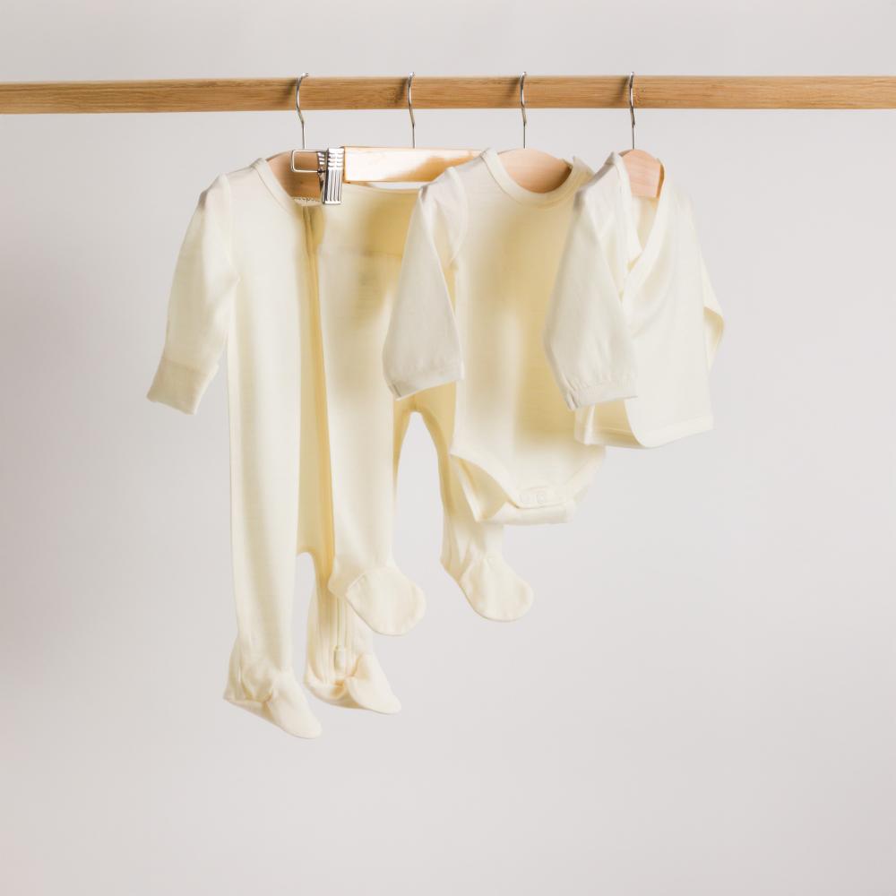 A set of cream-colored jersey merino baby items including footed zipsuit, footed leggings, bodysuit and kimono cardigan hanging on a wooden rack against a light background